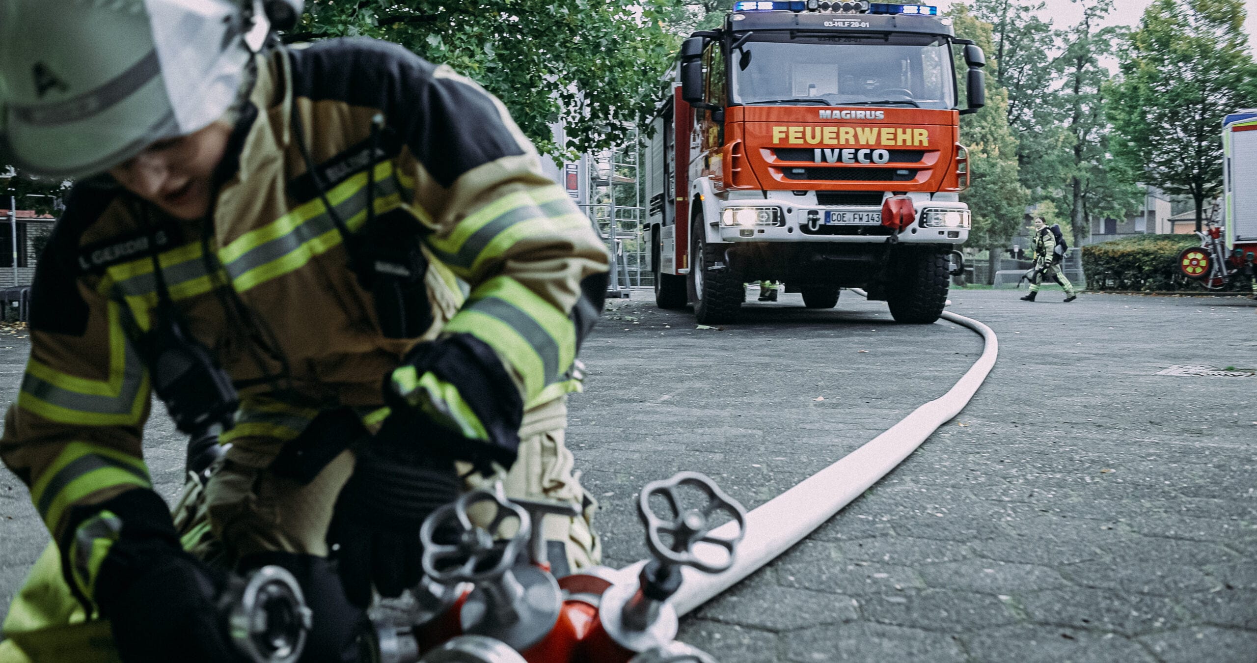 Feuerwehrfrau mit Schlauchleitung bei Einsatz auf der Straße, Löschfahrzeug im Hintergrund